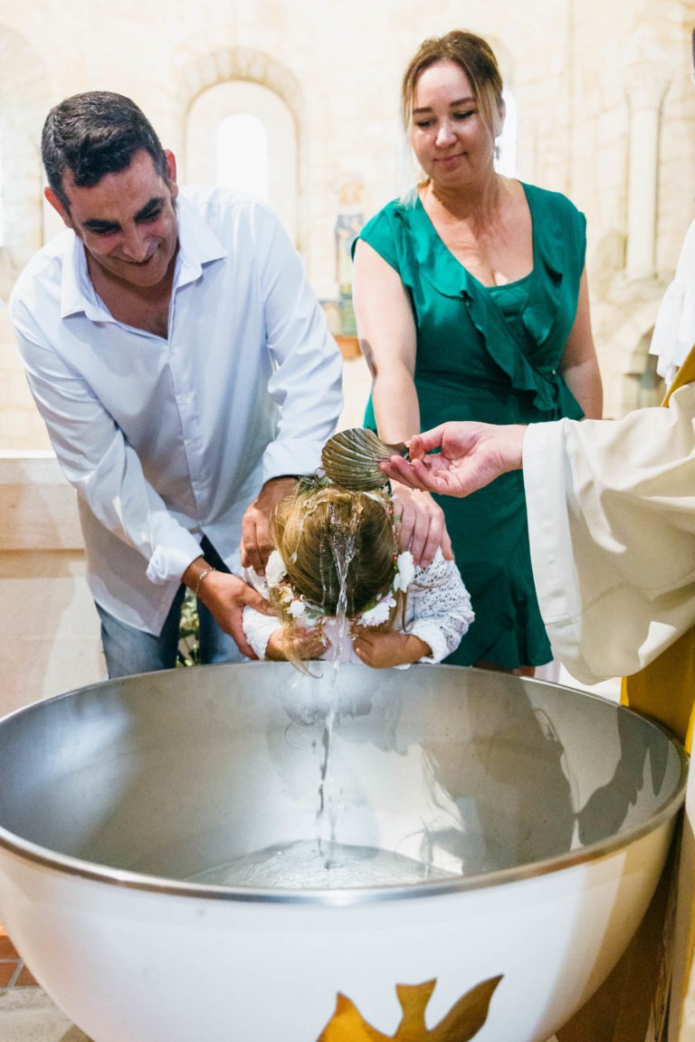 Un enfant, porté par un homme en chemise blanche, est baptisé dans une église. Une personne vêtue d'une robe couleur crème verse de l'eau sur la tête de l'enfant à partir d'une coquille Saint-Jacques. Une femme vêtue d'une robe verte se tient à proximité, observant le moment sacré capturé par le photographe de baptême près des grands fonts baptismaux ornés de symboles religieux.