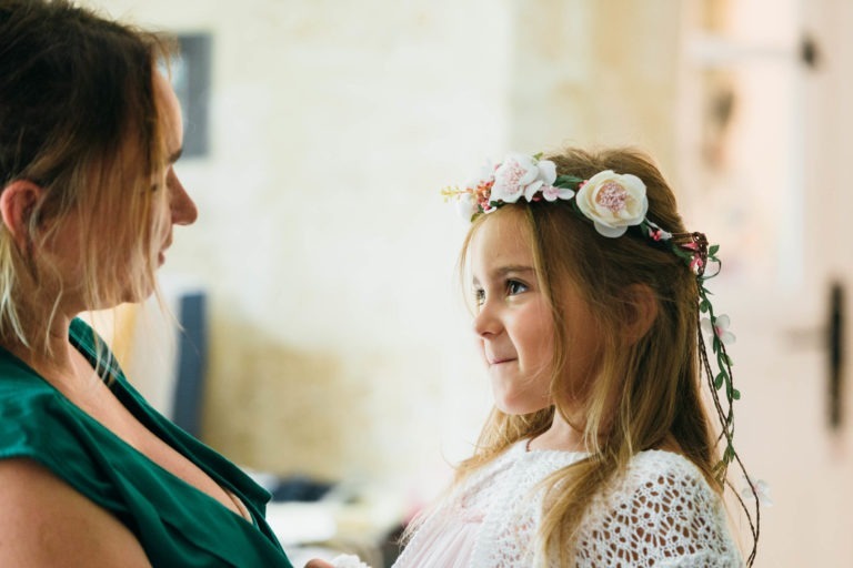 Une jeune fille vêtue d'une robe en dentelle blanche et d'une couronne de fleurs roses et blanches regarde une femme en vert qui lui sourit. Le flou doux de l'arrière-plan crée une atmosphère chaleureuse et intime, capturant un moment affectueux dans la pièce lumineuse - une scène parfaite pour tout photographe de baptême.