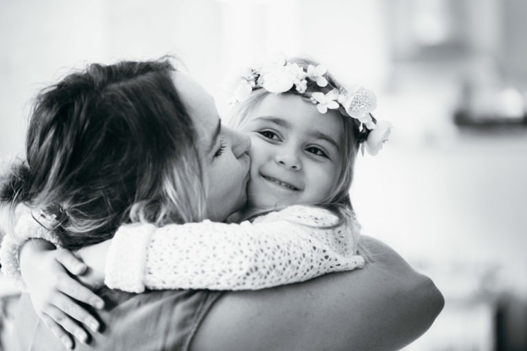 Une photo en noir et blanc prise par un photographe de baptême montre un adulte en train d'embrasser un jeune enfant souriant. L'enfant porte une couronne de fleurs et un haut en dentelle, ses bras enroulés autour du cou de l'adulte dans une étreinte chaleureuse. L'arrière-plan est flou, ce qui met en valeur le moment de tendresse entre les deux.