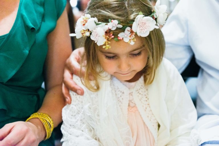 Une jeune fille aux cheveux châtain clair, coiffée d'une couronne de fleurs et d'un châle blanc, est assise entre deux adultes. Elle regarde vers le bas, l'air serein. La personne de gauche porte une robe verte et des bracelets jaunes, tandis que celle de droite est en blanc ; leur bras repose doucement autour de ses épaules pendant le photographe de baptême.