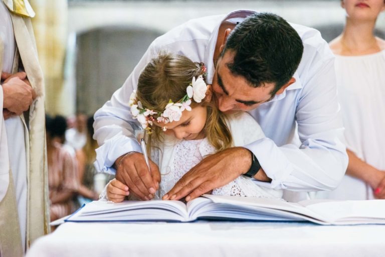 Un homme en chemise blanche se tient derrière une jeune fille portant un bandeau fleuri et l'aide à écrire dans un grand livre ouvert sur une table. Tous deux sont concentrés sur le livre. D'autres personnes sont floues à l'arrière-plan, suggérant une cérémonie ou une célébration en intérieur, probablement capturée par un photographe de baptême.