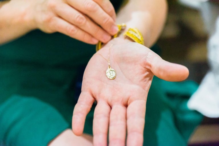 Une personne tient dans sa paume ouverte un bracelet en or avec une breloque ronde au motif complexe. L'autre main ajuste le bracelet. La personne, peut-être un photographe de baptême, porte un vêtement vert. L'accent est mis sur la breloque et la main, avec un arrière-plan flou.