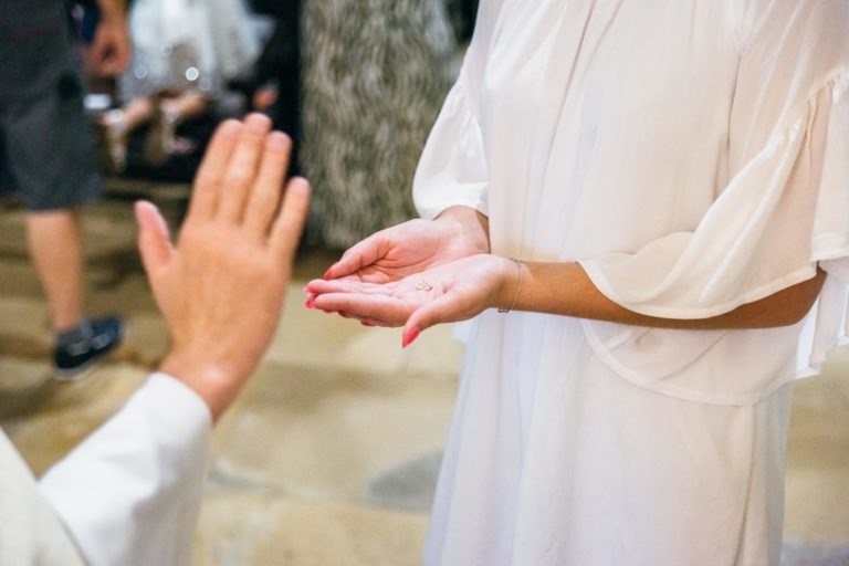 Une personne aux ongles vernis de rouge, vêtue d'un vêtement blanc, tient de minuscules objets dorés dans ses paumes ouvertes. Une autre personne, également vêtue de blanc, lève une main ouverte vers eux. La scène, capturée par un photographe de baptême, a un arrière-plan flou avec d'autres personnes visibles.