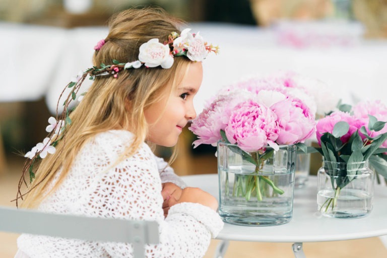 Une jeune fille aux longs cheveux blonds, vêtue d'une robe blanche en dentelle et d'un bandeau fleuri, est accoudée à une table. Elle sourit et regarde des pivoines roses dans des vases en verre posés sur la table. L'arrière-plan est flou, suggérant un décor intérieur capturé par un photographe de baptême.