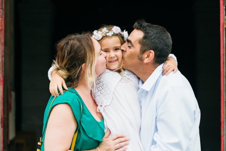 Une jeune fille en robe blanche et couronne de fleurs sourit tandis qu'elle est soulevée et embrassée sur les joues par une femme en haut vert à gauche et un homme en chemise blanche à droite. Parfaitement capturées par notre photographe de baptême, leurs expressions sont joyeuses et affectueuses sur le fond sombre.