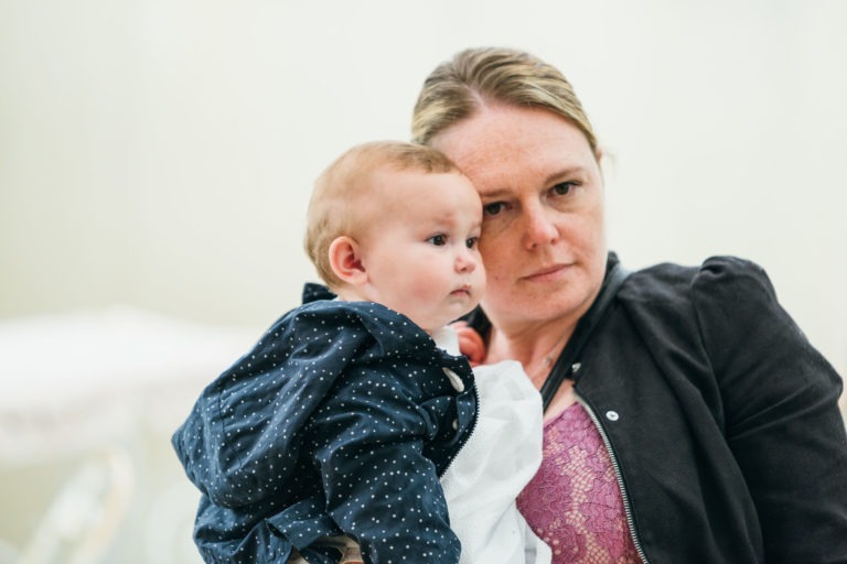 Une femme à la peau claire et aux cheveux châtain clair, vêtue d'une veste noire sur un haut violet, tient dans ses bras un bébé à la peau claire et aux cheveux châtain clair. Le bébé est habillé d'une tenue à pois bleu marine et a l'air attentif. Tous deux regardent dans la même direction avec des expressions neutres sur un fond neutre, comme s'ils étaient capturés par un photographe de baptême.