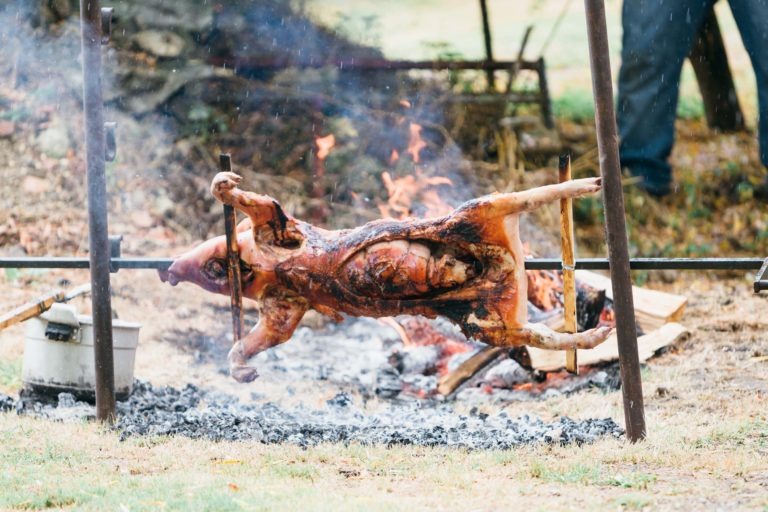 Un cochon entier est en train de rôtir sur une broche au-dessus d'un feu ouvert. La mise en scène comprend des supports métalliques qui maintiennent la broche, et le cochon est embroché et cuit uniformément. L'arrière-plan montre une zone herbeuse avec du bois et une photographie de baptême partiellement visible capturant l'atmosphère rustique renforcée par le feu et la fumée.