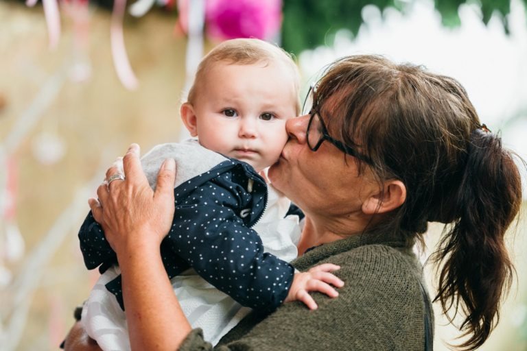 Un adulte âgé aux cheveux noirs et aux lunettes embrasse la joue d'un bébé qu'il tient dans ses bras. Le bébé, vêtu d'une veste bleue à pois blancs et d'un bavoir blanc, a les cheveux clairs. L'arrière-plan est flou, mettant en valeur la verdure et les décorations, capturant un moment tendre de photographie de baptême.