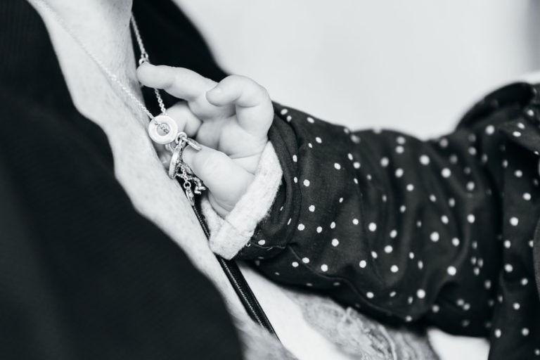 Gros plan en noir et blanc de la main d'un bébé saisissant un pendentif circulaire sur un collier porté par un adulte. Photographe de baptême capture le bébé vêtu d'une tenue à pois, en se concentrant sur l'interaction délicate entre la main du bébé et le collier, soulignant la tendresse du moment.