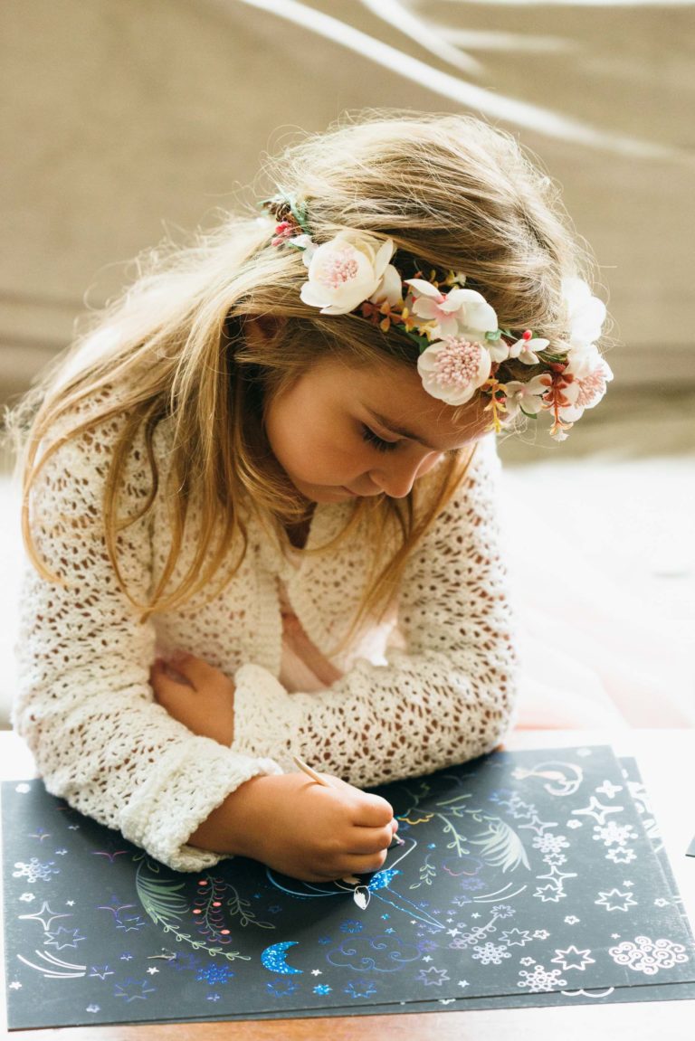 Une jeune fille aux cheveux châtain clair porte un pull blanc crocheté et un bandeau fleuri. Elle se concentre sur la création artistique avec un papier à gratter. Alors qu'elle utilise le stylet pour révéler des motifs scintillants, elle a l'impression d'être une photographe de baptême capturant la transformation magique de la couleur émergeant de l'obscurité.