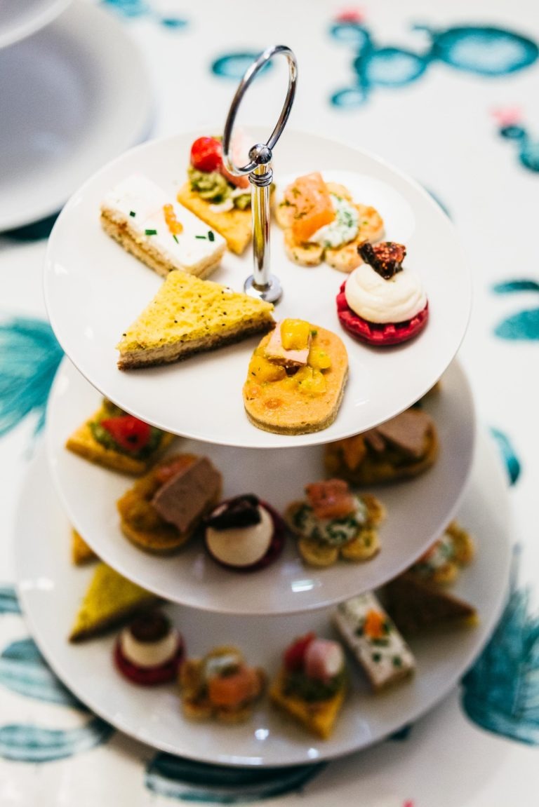 Un présentoir à desserts à trois niveaux abrite un assortiment d'amuse-gueules et d'amuse-gueules colorés, parfaits pour un photographe de baptême. Les éléments visibles comprennent des mini-sandwichs, des craquelins avec garniture, des canapés et des petites pâtisseries. L'arrière-plan présente une nappe blanche aux motifs bleus et verts.