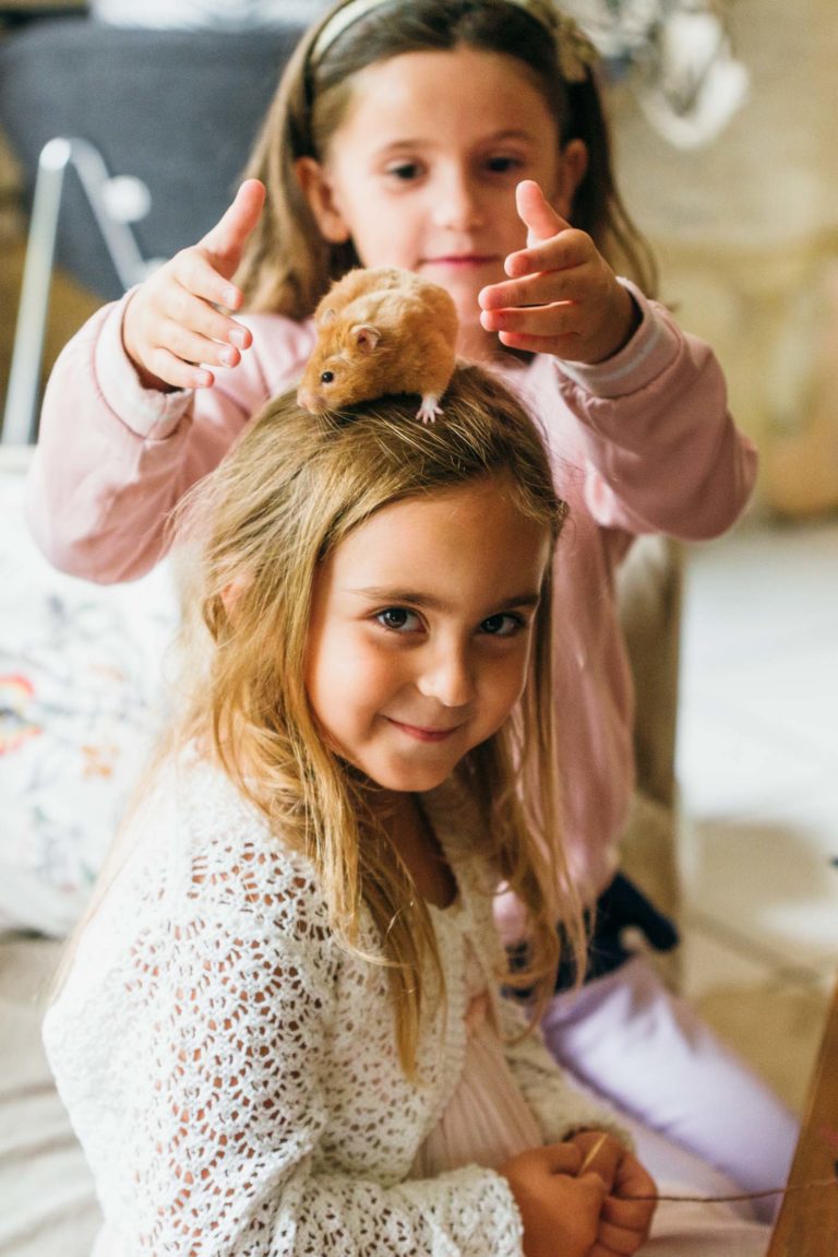 Deux jeunes filles jouent à l'intérieur. La fille au premier plan, vêtue d'un pull blanc crocheté, sourit tandis qu'un petit hamster est assis sur sa tête. La fille à l'arrière-plan, vêtue d'un pull rose, place soigneusement ses mains près du hamster, supervisant son placement avec une précision digne d'un photographe de baptême.