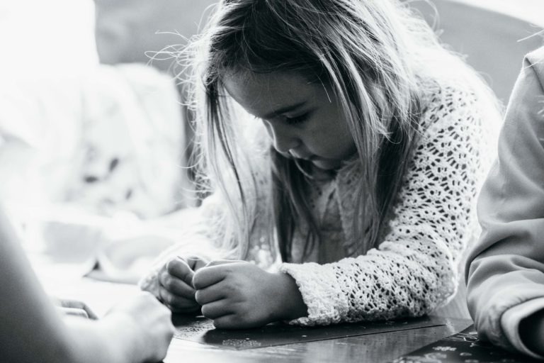 Image en noir et blanc d'une jeune fille concentrée sur une tâche. Elle a les cheveux longs et porte un pull en tricot. Ses mains manipulent de petits objets sur une surface plane, probablement en train de bricoler ou de faire un puzzle. Le décor semble être intérieur, rappelant une scène intime de photographe de baptême.