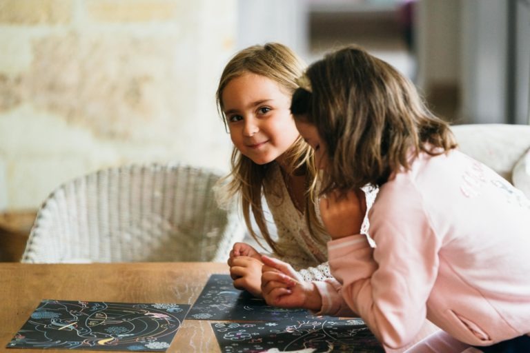 Deux jeunes filles sont assises à une table en bois et s'adonnent à des activités de dessin ou de coloriage sur du papier noir aux motifs colorés. L'une des filles aux longs cheveux blonds sourit à l'objectif, tandis que l'autre fille aux cheveux bruns courts, habillée en rose, se concentre sur l'œuvre d'art. C'est un moment serein que tout photographe de baptême aimerait capturer.