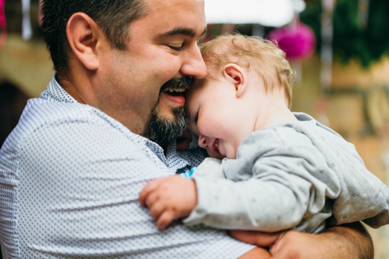 Un homme barbu aux cheveux noirs courts tient affectueusement un bébé souriant contre sa poitrine. L'homme, vêtu d'une chemise blanche à motifs, semble heureux et satisfait alors qu'il embrasse le bébé dans une tenue gris clair. Capturée par un photographe de baptême, la scène respire la chaleur et la joie.