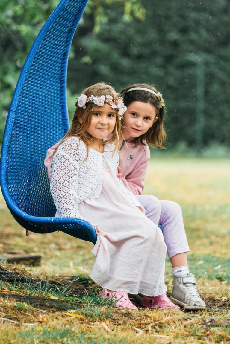 Deux jeunes filles sont assises sur une balançoire bleue à l'extérieur. La fille de gauche porte une robe en dentelle blanche, des chaussures roses et un bandeau fleuri, tandis que la fille de droite est habillée d'un haut rose et d'un pantalon violet pâle avec des baskets blanches. Elles sont entourées de verdure par une journée légèrement pluvieuse, parfaite pour une séance de photographe de baptême.