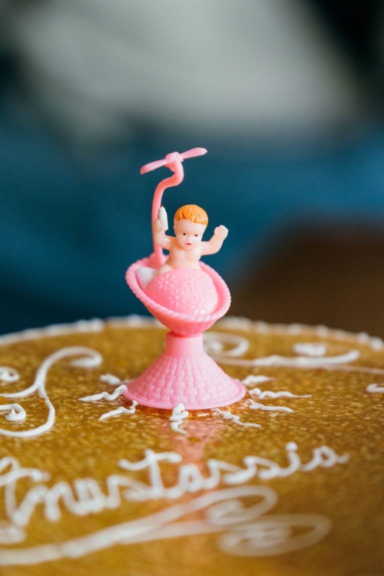 Une figurine de bébé en plastique aux cheveux orange et à la couche rose se tient debout, les bras levés, à l'intérieur d'un objet creux, rose, en forme de cône. La figurine est posée sur une surface dorée et scintillante ornée d'une écriture cursive blanche, idéale pour un photographe de baptême. Arrière-plan flou.