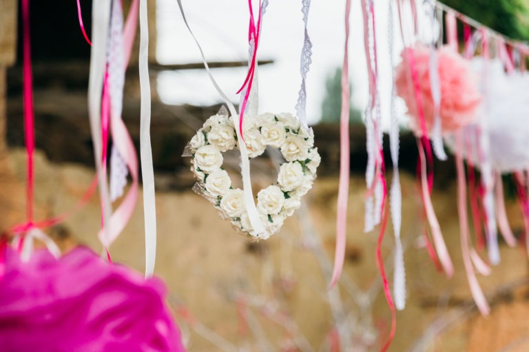 Une couronne en forme de cœur composée de roses blanches est suspendue entre divers rubans et décorations. Les rubans, dans des tons de blanc, de rose et de rouge, ajoutent une touche festive et romantique. L'arrière-plan est flou, mais il semble s'agir d'un décor extérieur avec des éléments naturels, parfait pour un photographe de baptême pour capturer des moments spéciaux.