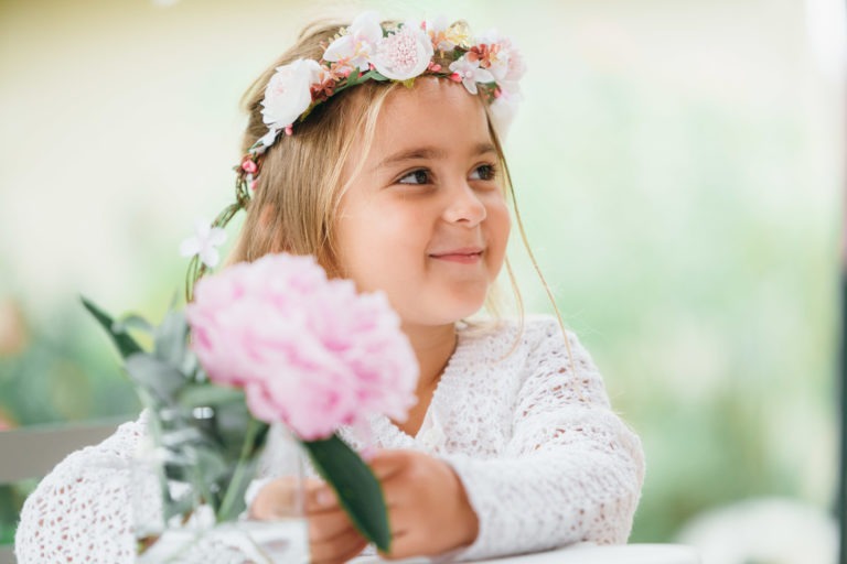 Une jeune fille aux cheveux blonds, souriant doucement, est parée d'une couronne de fleurs blanches et roses. Elle porte un haut en tricot blanc et tient un vase en verre avec une fleur rose. L'arrière-plan est légèrement flouté avec des teintes vertes suggérant un décor extérieur, parfait pour un photographe de baptême.