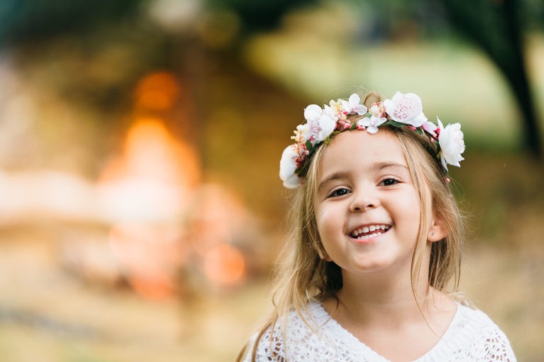 Une jeune fille aux longs cheveux blonds sourit chaleureusement. Elle porte un bandeau à fleurs blanc et un haut en dentelle blanche. L'arrière-plan est flou, avec une lumière douce et chaleureuse qui suggère un décor extérieur chaleureux, peut-être près d'un feu. L'ambiance est joyeuse et sereine, parfaite pour une séance de photographe de baptême.