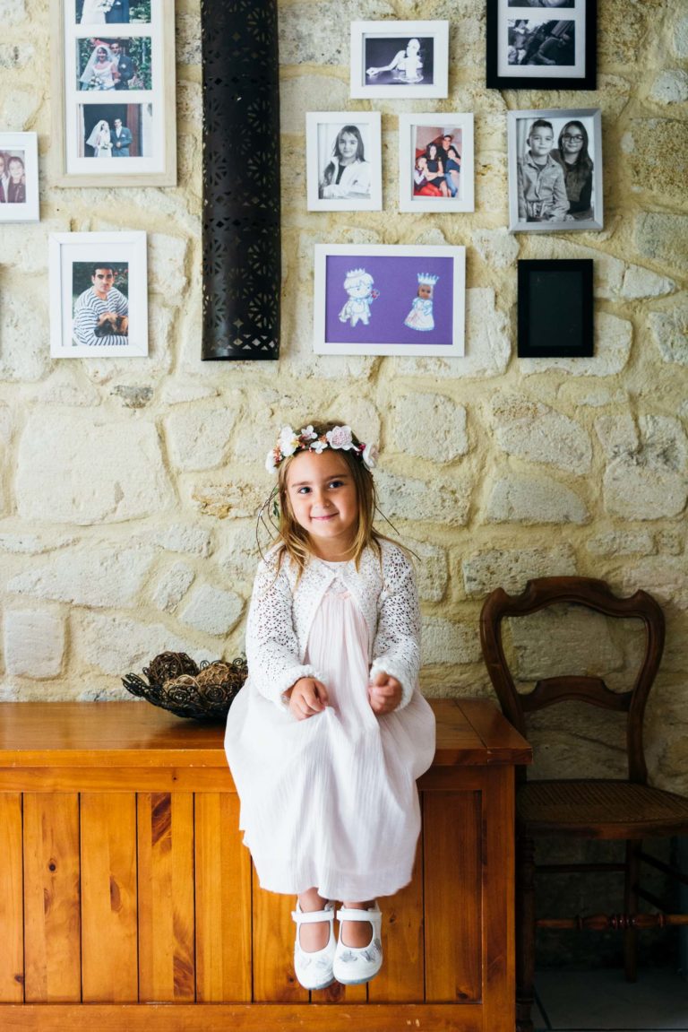 Une jeune fille vêtue d'une robe blanche, de chaussures blanches et d'une couronne de fleurs est assise sur un coffre en bois dans une pièce rustique au mur de pierre. Au-dessus d'elle, Photographe de baptême capture l'instant tandis qu'une galerie de photos encadrées et d'œuvres d'art ajoute du charme. À proximité, une chaise en bois et un bol décoratif complètent la scène.