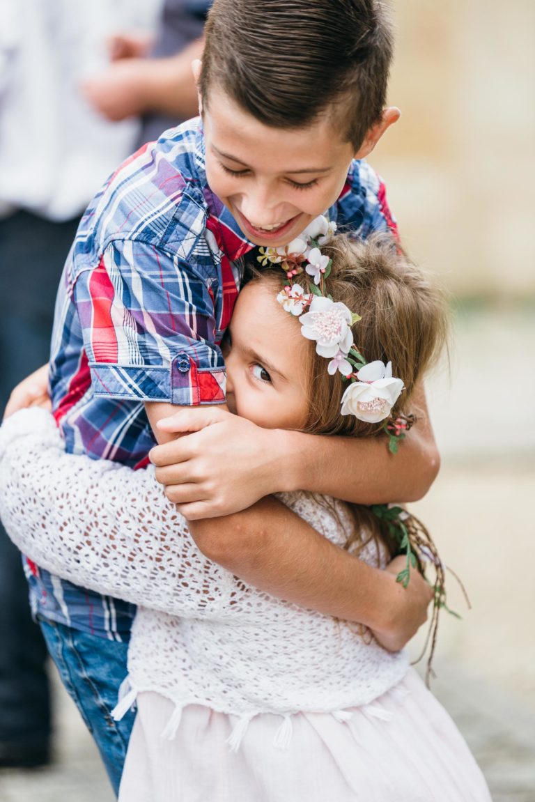 Un jeune garçon en chemise à carreaux serre dans ses bras une petite fille avec un bandeau fleuri et un pull en dentelle blanche. La fille sourit et regarde le garçon, capturant un moment de joie et d'affection. Le photographe de baptême a habilement flouté l'arrière-plan pour garder l'accent sur les expressions et l'étreinte des enfants.