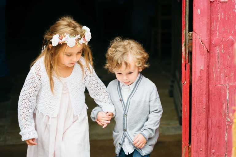 Une jeune fille et un garçon se tiennent la main en marchant. La fille, vêtue d'une robe blanche, d'un bandeau fleuri et d'un cardigan en tricot blanc, guide le garçon, qui porte un cardigan gris sur une chemise rayée. Ils se tiennent à côté d'une porte rouge et le photographe de baptême capture ce moment tendre alors que le garçon regarde vers le bas.