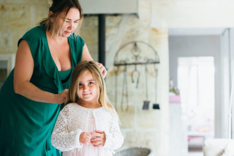 Un photographe de baptême capture un moment tendre alors qu'une femme en robe verte se tient derrière une jeune fille en train de coiffer ses cheveux blonds. La fille porte un cardigan en dentelle blanche sur une robe rose pâle et sourit doucement. Elles sont à l'intérieur, avec un arrière-plan rustique et doucement éclairé, avec des murs de couleur claire et divers objets suspendus.