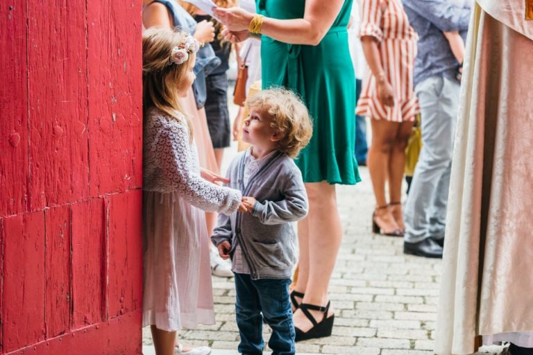 Deux jeunes enfants, une fille et un garçon, se tiennent près d'une surface en bois rouge. La fille porte une robe lavande et un bandeau fleuri, tandis que le garçon a les cheveux blonds bouclés et porte un cardigan gris sur une chemise bleue et un jean. Ils semblent engagés dans une conversation tandis que des adultes les entourent en arrière-plan, capturant des moments comme ceux que chérit tout photographe de baptême.