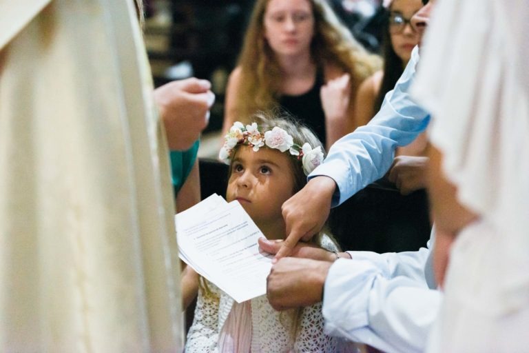 Une jeune fille portant une couronne de fleurs lève les yeux tout en tenant des papiers, entourée d'adultes aux vêtements clairs qui pointent les documents du doigt. En arrière-plan, plusieurs personnes aux visages flous sont assises, concentrées sur la scène. Le photographe de baptême capture magnifiquement le cadre cérémoniel ou formel.