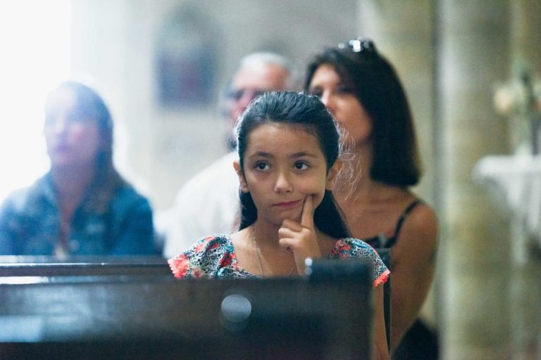 Une jeune fille à l'expression pensive, le menton posé sur sa main, est assise dans une église. Elle porte un haut coloré et a les cheveux noirs attachés en arrière. Derrière elle, plusieurs adultes sont assis, en partie flous. Le décor semble calme et serein avec un éclairage doux, parfait pour tout photographe de baptême souhaitant capturer l'instant.