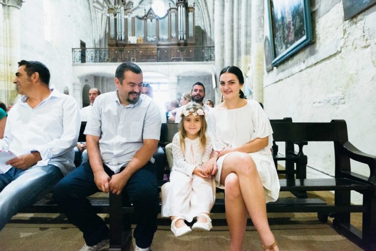 Une jeune fille portant une couronne de fleurs est assise entre un homme et une femme sur un banc d'église. La jeune fille sourit à l'objectif tout en tenant la main de la femme. D'autres personnes sont assises le long du banc et d'autres sont visibles à l'arrière-plan. L'intérieur lumineux de l'église, parfait pour un photographe de baptême, présente des pierres détaillées et un grand orgue en vue.