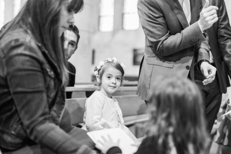 Photographie en noir et blanc d'une jeune fille portant une couronne de fleurs, assise sur un banc parmi des adultes. Elle regarde un autre enfant au premier plan, tandis que les adultes interagissent. Le décor, une église avec des vitraux en arrière-plan, évoque un baptême mémorable à Bordeaux.