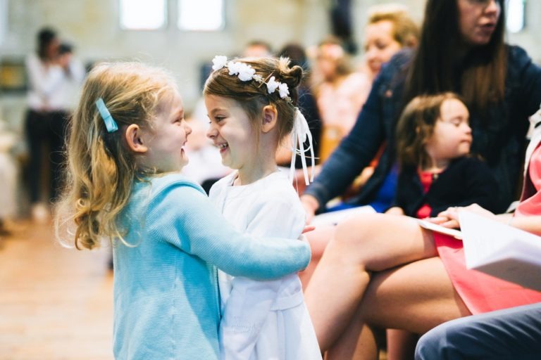 Deux jeunes filles, l'une vêtue d'un cardigan bleu clair et l'autre d'une robe blanche avec un bandeau fleuri, s'embrassent et se sourient. Elles sont dans une salle remplie d'adultes et d'enfants assis, qui sourient tous et les regardent. L'ambiance est joyeuse et chaleureuse lors de ce baptême mémorable à Bordeaux.