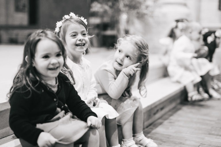 Photographie en noir et blanc de trois jeunes filles assises sur une marche. Elles sourient toutes et portent des robes, avec une fille au centre qui porte un bandeau fleuri. L'arrière-plan montre un environnement intérieur flou avec d'autres enfants flous au loin, capturant un baptême mémorable à Bordeaux.