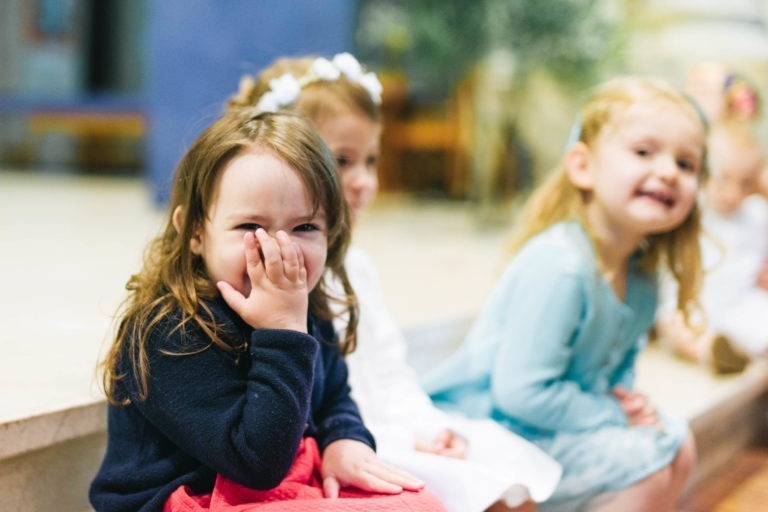 Trois jeunes enfants sont assis en rang. L'enfant de gauche, aux longs cheveux bruns, se couvre la bouche et rit. L'enfant du milieu a les cheveux bruns courts et regarde légèrement ailleurs. L'enfant de droite, aux longs cheveux blonds, sourit à la caméra. Tous portent des vêtements colorés, rappelant un baptême mémorable à Bordeaux.