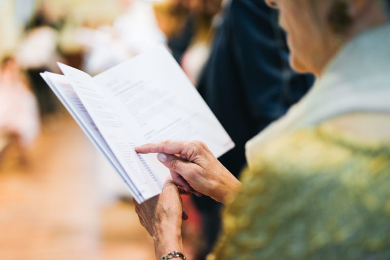 Une femme, vêtue d'un vêtement jaune-vert, tient dans ses mains une page d'un livret ou d'un programme et la désigne du doigt. L'arrière-plan est flou, laissant apparaître d'autres personnes et créant une ambiance douce et chaleureuse. Son attention semble se porter sur le texte, lisant ou recherchant peut-être une section spécifique liée au Baptême mémorable à Bordeaux.