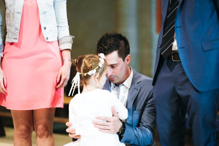 Un homme en costume est à genoux et tient dans ses bras une jeune fille portant un bandeau à fleurs blanches et une robe blanche. À côté d'eux, une femme portant une robe rose et une veste en jean est partiellement visible. Une autre personne en costume se tient à droite. La scène, qui capture un baptême mémorable à Bordeaux, semble se dérouler à l'intérieur d'un bâtiment avec un éclairage tamisé.