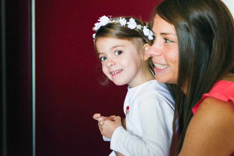 Une jeune fille avec un bandeau à fleurs blanc et de longs cheveux bruns sourit en regardant légèrement sur le côté. Elle porte un haut blanc et a les mains jointes. À côté d'elle, une femme adulte aux longs cheveux bruns portant un haut rose sourit également en regardant vers l'avant, célébrant un baptême mémorable à Bordeaux sur un fond rouge foncé.