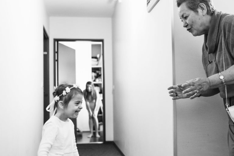 Une photographie en noir et blanc montre une jeune fille souriante marchant dans un couloir. Elle porte un bandeau orné de fleurs et s'approche d'une femme plus âgée qui applaudit et sourit. En arrière-plan, on voit un autre enfant debout dans l'embrasure d'une porte. La scène paraît joyeuse et accueillante, rappelant un baptême mémorable à Bordeaux.