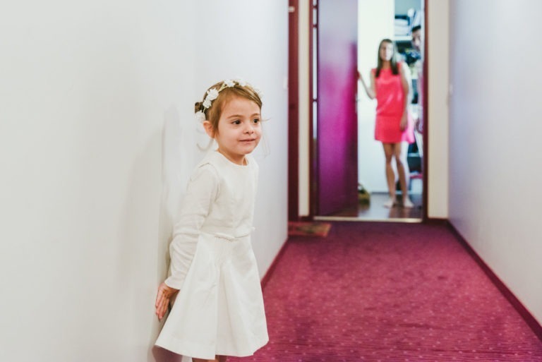 Une jeune fille en robe blanche et coiffe fleurie se tient dans un couloir au tapis rose et aux murs blancs. Elle regarde vers sa gauche. En arrière-plan, une femme en robe rose franchit une porte, capturant l'essence d'un baptême mémorable inoubliable à Bordeaux.