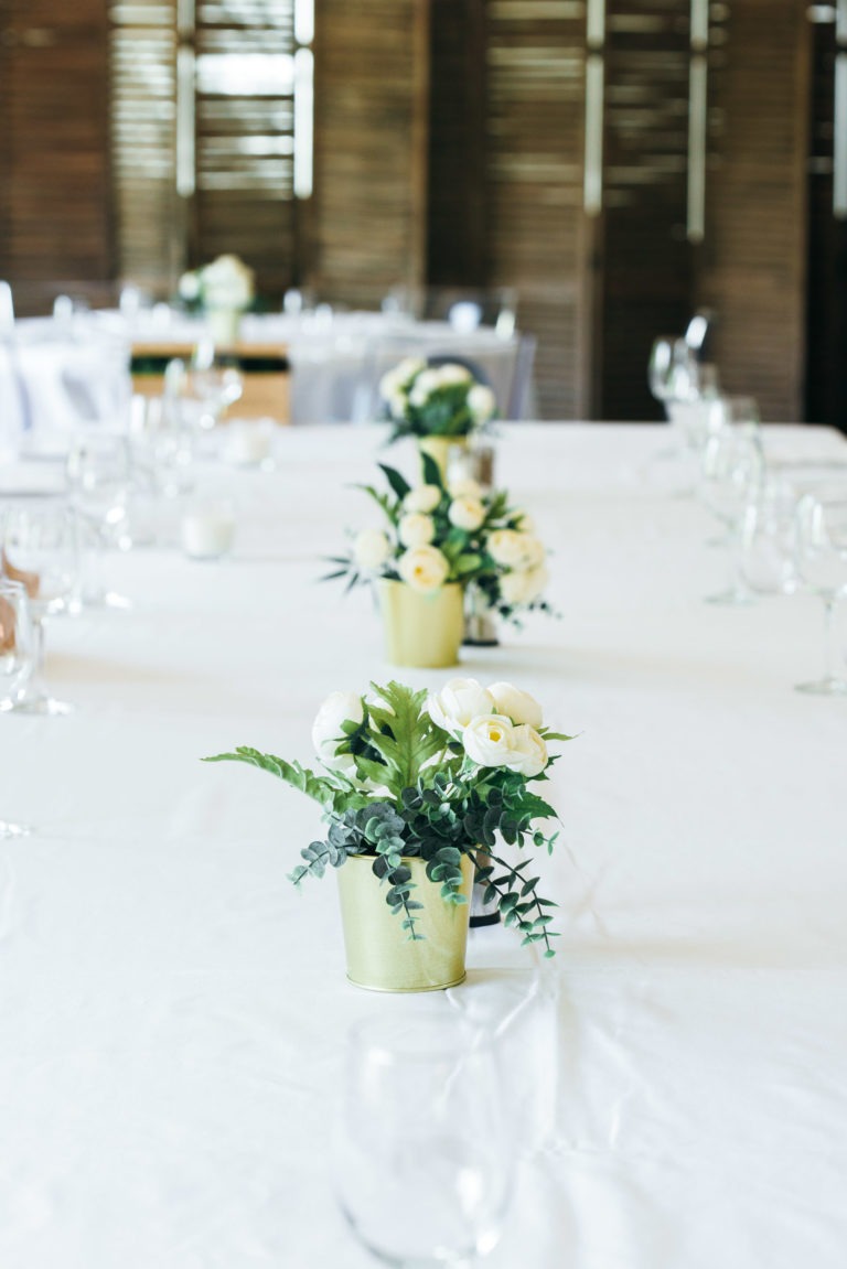 Une longue table recouverte d'une nappe blanche est dressée avec de la verrerie et décorée de petites compositions florales dans des pots dorés. Les compositions sont composées de roses blanches et de verdure. Le fond met en valeur des boiseries aux lumières tamisées, créant une ambiance élégante pour un baptême mémorable à Bordeaux.