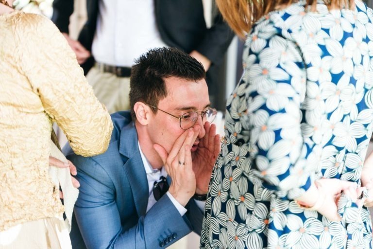 Un homme en costume bleu est accroupi, penché vers le ventre d'une femme enceinte et parle avec les mains en coupe autour de sa bouche. La femme, vêtue d'une robe à motifs floraux, sourit chaleureusement. D'autres personnes en tenue de soirée sont à l'arrière-plan, légèrement floues, célébrant un baptême mémorable à Bordeaux.