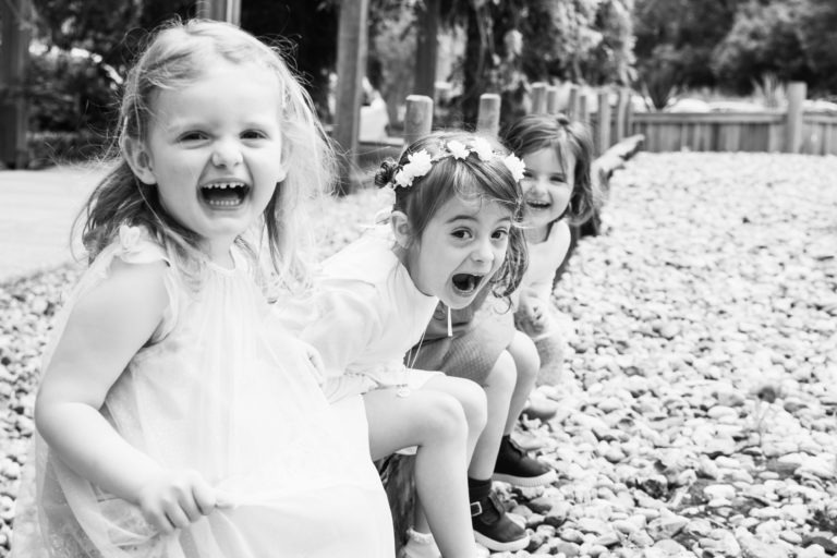 Trois jeunes enfants sont assis sur une poutre en bois près d'un sol recouvert de galets, ils rient et ont l'air excités. Une fille à gauche porte une robe légère, l'enfant du milieu porte une couronne de fleurs et pointe vers l'avant, et la fille à droite se penche joyeusement. La scène capture un baptême mémorable à Bordeaux avec des arbres derrière eux.