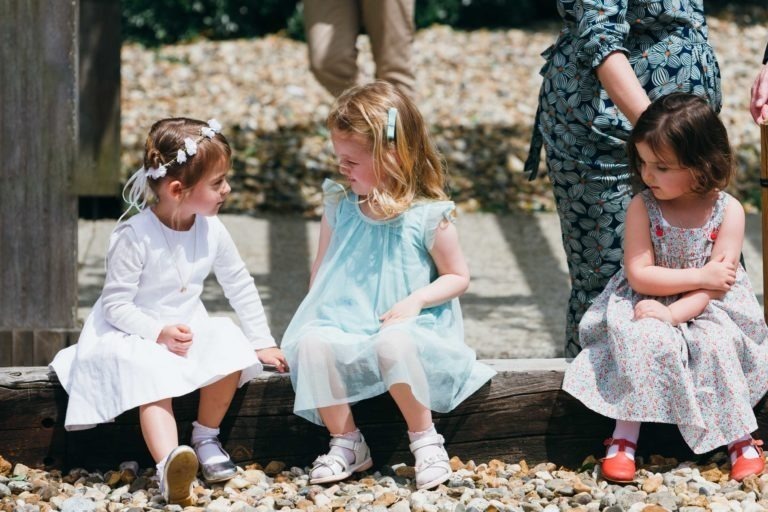 Trois jeunes filles assises sur une poutre en bois dans une zone jonchée de galets lors d'un baptême mémorable à Bordeaux. La fille de gauche est habillée en blanc avec un bandeau fleuri, celle du milieu porte une robe bleu pastel et celle de droite porte une robe fleurie avec des chaussures rouges. Deux adultes se tiennent à l'arrière-plan.