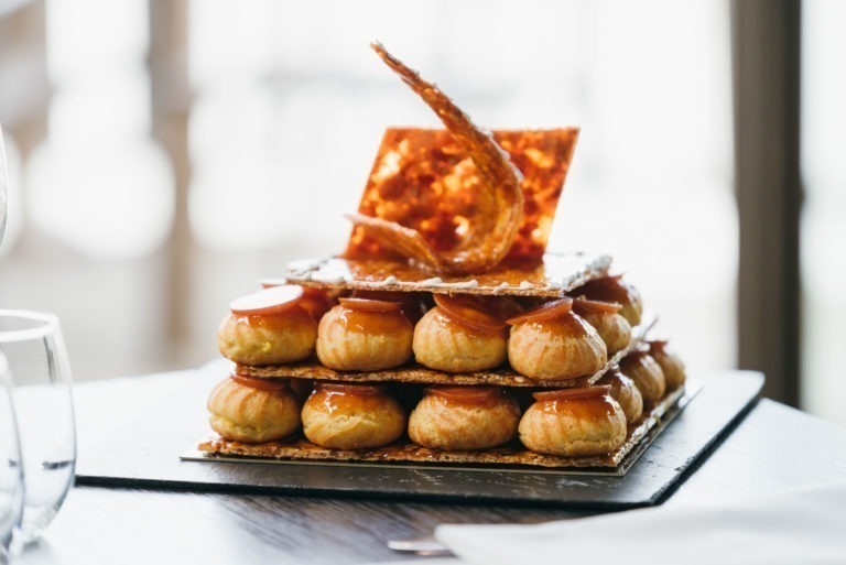 Un dessert soigneusement élaboré orne la table, célébrant un baptême mémorable à Bordeaux. Il se compose de deux couches de boules de pâtisserie nappées de caramel, posées sur de fines gaufrettes. Un morceau de caramel décoratif ajoute de la hauteur et de l'élégance. Deux verres à vin vides sont visibles au premier plan à gauche.