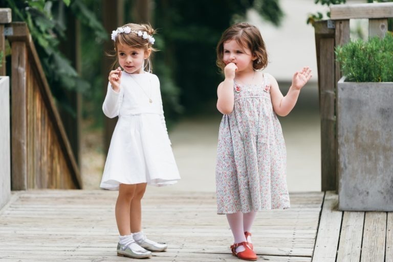 Deux jeunes filles se tiennent debout sur une terrasse en bois au milieu d'une végétation luxuriante. La fille de gauche, vêtue d'une robe blanche avec un bandeau fleuri et des chaussures argentées, tient un petit objet. La fille de droite, vêtue d'une robe à motifs et de chaussures rouges, se tient debout, une main près de la bouche et l'autre levée pendant ce baptême mémorable à Bordeaux.