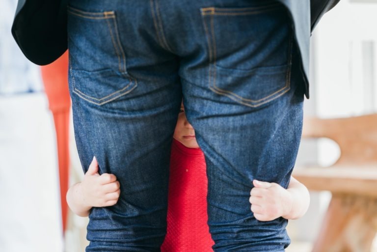 Un petit enfant en chemise rouge regarde à travers les jambes d'un adulte, tenant son jean bleu. L'adulte, vêtu d'une veste noire, se tient debout au milieu de la scène animée de ce qui semble être un baptême mémorable à Bordeaux, avec un arrière-plan flou qui comprend des personnes et des meubles.