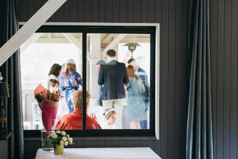 Un groupe de personnes est réuni et échange sur un balcon, vu à travers une grande fenêtre depuis une pièce intérieure. A l'intérieur, une table est ornée d'un bouquet de fleurs et d'une plante en pot. Les murs en bois peints en gris et le rideau à droite encadrent cette scène conviviale et animée qui rappelle un baptême mémorable à Bordeaux.