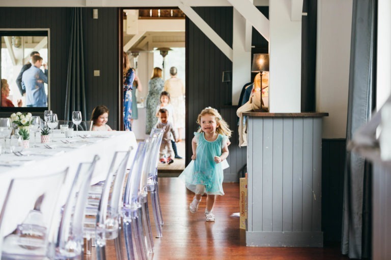 Une jeune fille en robe bleu clair court vers la caméra avec enthousiasme, souriante, dans une pièce bien éclairée au sol en bois. La pièce, décorée pour un baptême mémorable à Bordeaux, comporte une table avec des verres et des couverts à gauche. Plusieurs autres enfants et adultes sont visibles à l'arrière-plan.